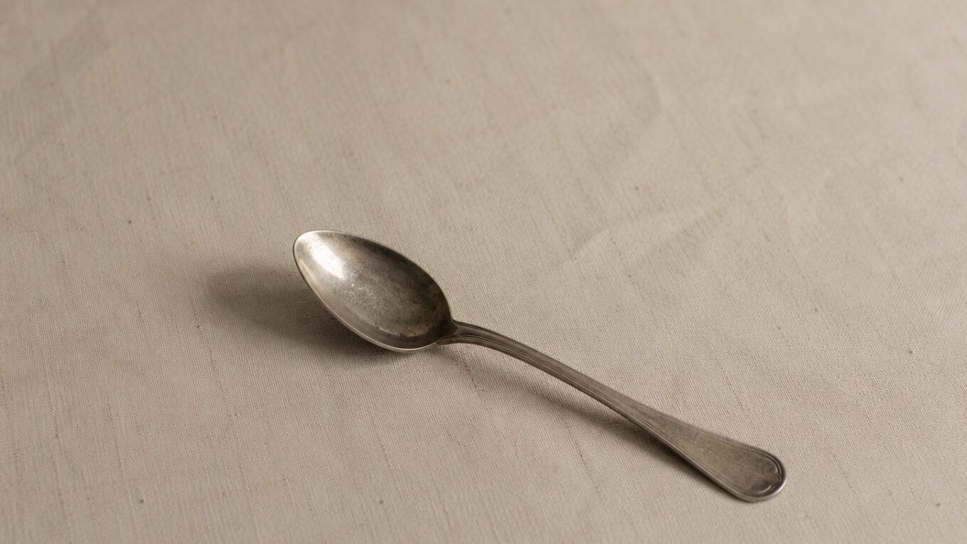 A single worn teaspoon rests on a muted linen surface, lit softly from one side with generous empty space around it.